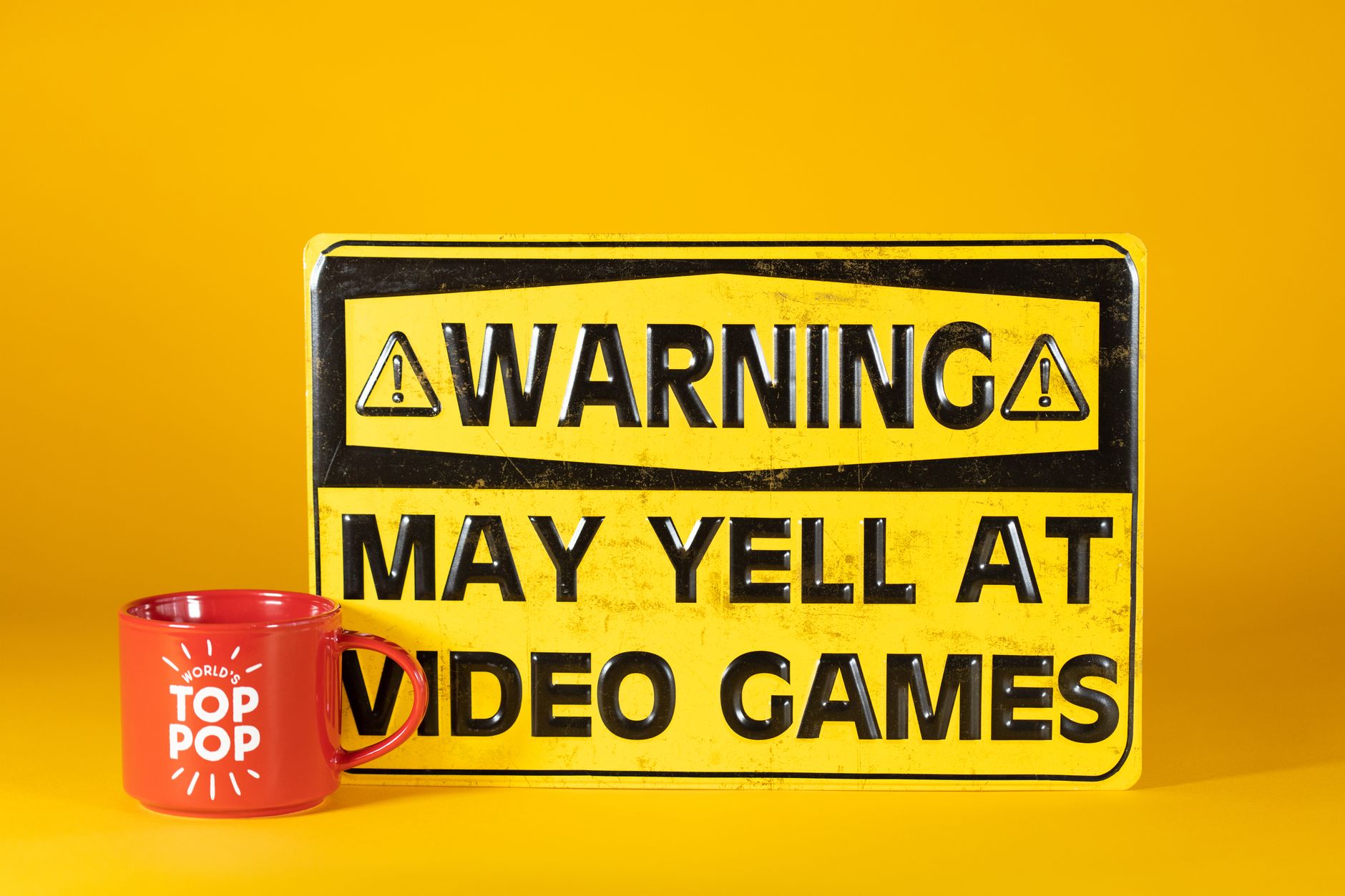 Papan peringatan lucu tentang video game dengan mug merah di latar belakang kuning cerah.